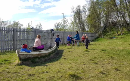Amaliahaugen barnehage uteområde Barn og voksne på uteområdet med trebåt, grønt gress og høye trær en solrik sommerdag