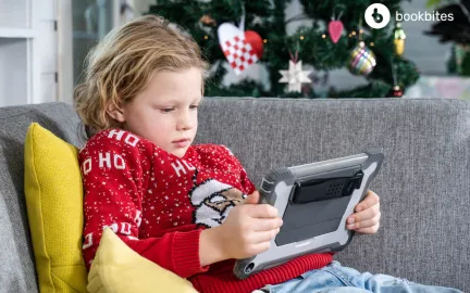 Barn i r&oslash;d genser med nisse p&aring; ser p&aring; et nettbrett. I bakgrunnen st&aring;r et juletre. Foto.