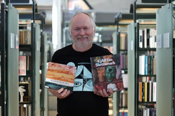 Øystein 150-årsutstilling Mann i svart skjorte med lyseblått trykk holder opp to bøker og smiler mot kamera. Foto.