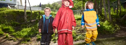 Tre barn med regnt&oslash;y i Fjellvegen barnehage en sommerdag med gr&oslash;nt gress og skog