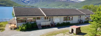 Lakselvbukt barnehage sett utenfra med lekeomr&aring;de, hav og fjell en str&aring;lende sommerdag