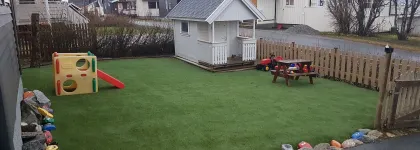 Lekeomr&aring;det i hagen med dukkehus, sklie, benk og bord p&aring; gr&oslash;nt gress en sommerdag